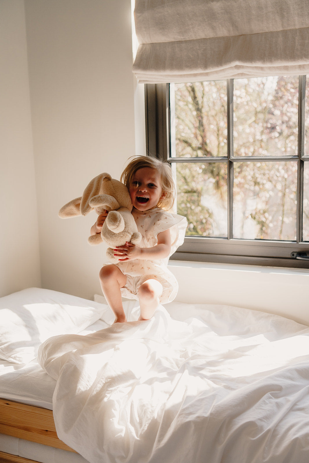 Klein meisje springt op bed met zachte beige knuffelbeer Boncoeur van Atelier Bonheur, ideaal geboortecadeau en eerste knuffel voor baby’s en kinderen.