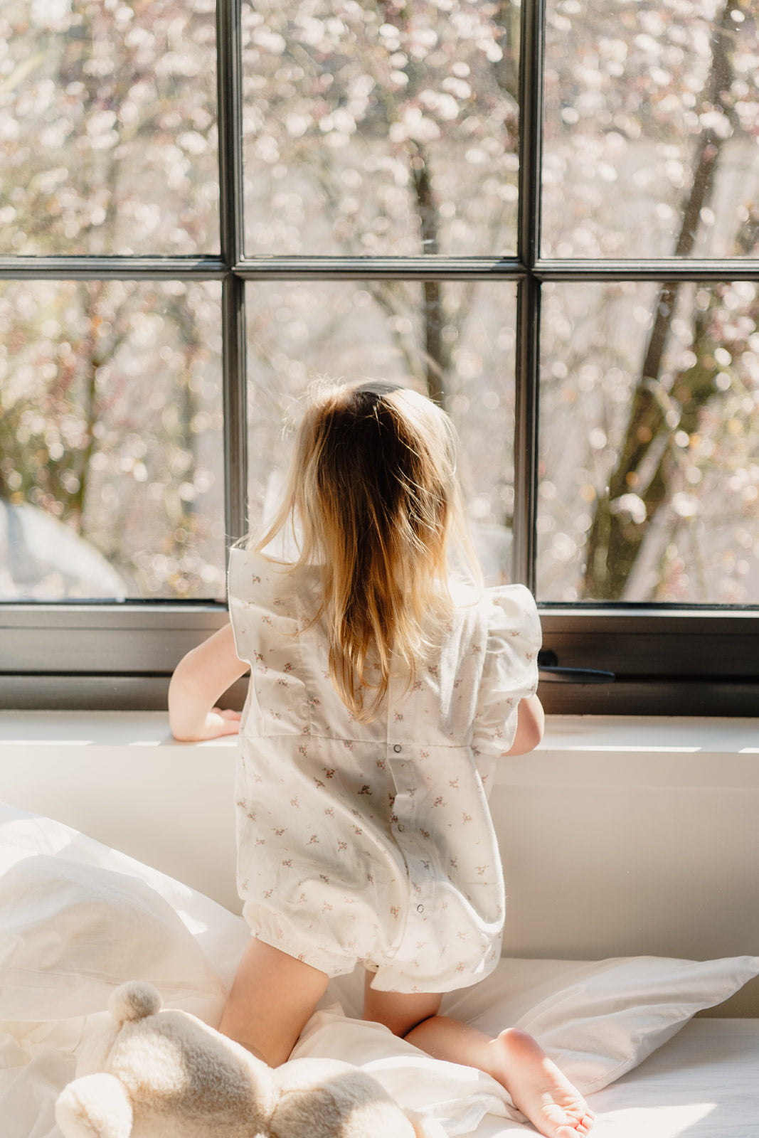 Meisje kijkt dromerig uit het raam in een katoenen bloemenpyjama uit de Cozy Dawn collectie van Atelier Bonheur, ontworpen voor rustige momenten thuis.

